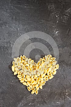 Raw macaroni - pasta Conchiglie. In the middle of the empty space in the shape of a heart on a dark concrete background.