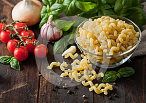 Raw maccheroni elbows pasta in glass bowl with oil and garlic  basil plant and tomatoes with pepper on wooden background