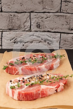 raw marbled meat pork steak with thyme and seasoning on brown parchment paper on a black table against a gray brick wall backgrou