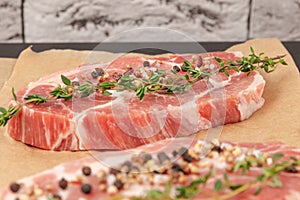 raw marbled meat pork steak with thyme and seasoning on brown parchment paper on a black table against a gray brick wall backgrou