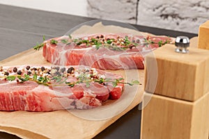 raw marbled meat pork steak with thyme and seasoning on brown parchment paper on a black table against a gray brick wall backgrou