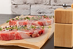 raw marbled meat pork steak with thyme and seasoning on brown parchment paper on a black table against a gray brick wall backgrou