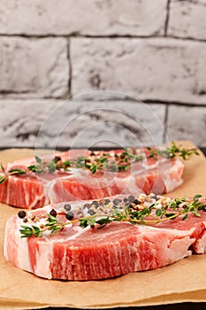 Raw marbled meat pork steak with thyme and seasoning on brown parchment paper on a black table against a gray brick wall backgroun