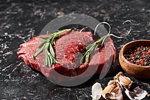 Raw meat steaks with rosemary twigs near small bowl with peppercorns and garlic on black marble surface.