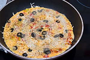 Raw omelet in a round frying pan, top view. Eggs, milk, cheese, olives, tomatoes, spices. Ready for further cooking.