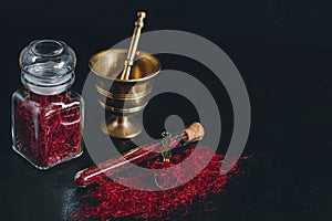 Raw organic red dried saffron spice on wooden background in vintage metal brass mortar with pestle, glass jar and tube