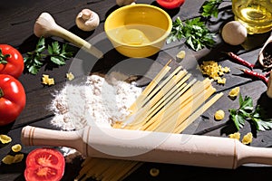 Raw pasta, tomatoes,mushrooms, flour and eggs on black wooden table background, top view.
