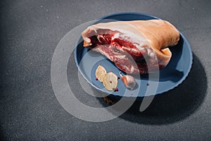 Raw pork knuckle on blue plate, seasonings, garlic, pepper set, bay leaf on grey background, close up. Cooking of meat dish, meal