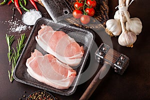 Raw pork loin chops in plastic tray with salt and pepper and vintage meat hatchets and hammer on rusty background.Red pepper,