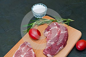 Raw pork steak, rosemary, tomatoes and salt on wooden cutting board, dark background, top view and side view