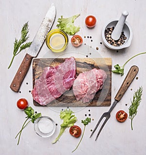 Raw Pork steak with vegetables and herbs, meat knife and fork, on a cutting board wooden rustic background top view close up