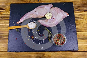 Raw rabbit legs with spices on a black slate cutting board on wooden table. Top view