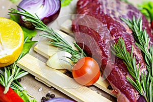 Raw red beef meat with green rosemary and fresh vegetables on light wooden cutting board background.