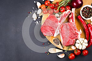 Raw ribeye beef steaks with seasonings and vegetables over dark concrete background with copy space for text