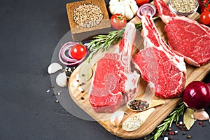 Raw ribeye beef steaks with seasonings and vegetables on wooden cutting board over dark concrete background