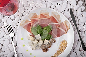A raw and salted ham, blue cheese and basil on a white plate. A glass of red wine and meat dish on the white stone