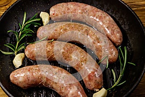 Raw sausages ready for preparation with rosemary, garlic and spices in cast iron grill frying pan on wooden table