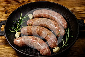 Raw sausages ready for preparation with rosemary, garlic and spices in cast iron grill frying pan on wooden table