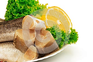 Raw slices of fish hake, pollock on the plate with lemon and leaves of salad lettuce on white background