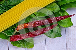 Raw spaghetti and hot red pepper stand on spinach leaves on a white wooden table