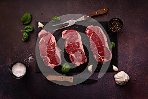 Raw striploin steaks on a black slate board with seasoning and herbs.