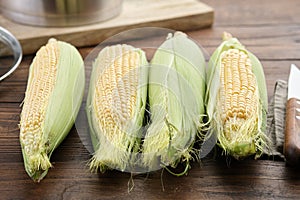 Raw sweet corn cobs for cooking and kitchen knife on table, cooking pot on background