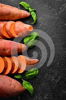 Raw sweet potatoes and basil on a black stone background. Top view.