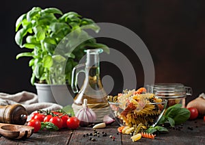 Raw tricolor fusilli pasta in glass bowl with oil and garlic, basil plant and tomatoes on wooden background