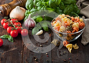 Raw tricolore trottole wheat pasta in glass bowl with oil and garlic, basil plant and tomatoes with pepper and linen towel on