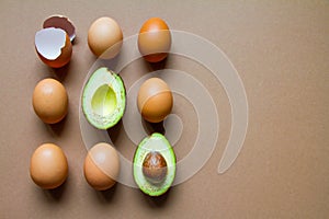 Raw two halves of avocado, few chicken eggs and eggshell on a brown background, flat, top view