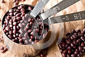 Raw, uncooked, dried adzuki red mung beans in metal measurement cups on brown packing paper background