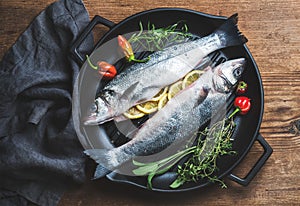 Raw uncooked seabass fish with lemon slices, herbs and spices on black grilling iron pan over rustic wooden background