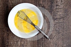 Raw whisked eggs in white bowl with metal whisk, wood table