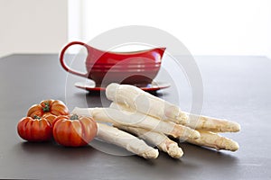 Raw white asparagus spears with pisanello tomatoes and a red gravy boat. Fresh vegetables on black table background