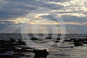 Rays of light phenomena over the sea horizon. Sunset background with sky clouds on a rocky beach.