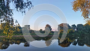 The rays of the sun setting over the river stain the willow branches bent over the water. The sky and trees are reflected in the c