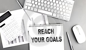 REACH YOUR GOALS text written on notebook on grey background with chart and keyboard, business concept