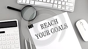 REACH YOUR GOALS written on paper with office tools and keyboard on the grey background