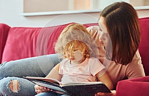 Reading big book. Young mother with her little daughter in casual clothes together indoors at home