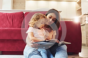 Reading big book. Young mother with her little daughter in casual clothes together indoors at home