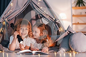 Reading book and using flashlight. Two little girls is in the tent in domestic room together