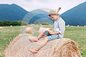 Reading boy sits over the haystack roll on the mountain field in