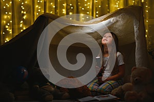 Reading and family games in children`s tent. Little girl with books and flashlight before going to bed