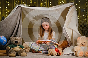 Reading and family games in children`s tent. Little girl with books and flashlight before going to bed