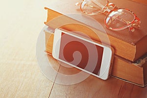 Reading glasses, stack of old books and smartphone over wooden table, retro filtered image