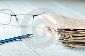 Reading the morning news. A stack of newspapers, glasses and a pencil on a wooden table lit by the morning sun