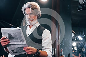 Reading newspaper. Stylish modern senior man with gray hair and beard is indoors