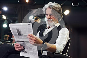 Reading newspaper. Stylish modern senior man with gray hair and beard is indoors