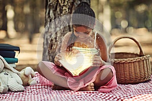 Reading is one way to nurture a childs curiosity. a little girl reading a book with glowing pages in the woods.