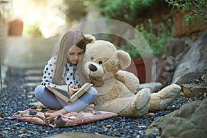 Reading their favourite story. a little girl reading a book with her teddy bear beside her.